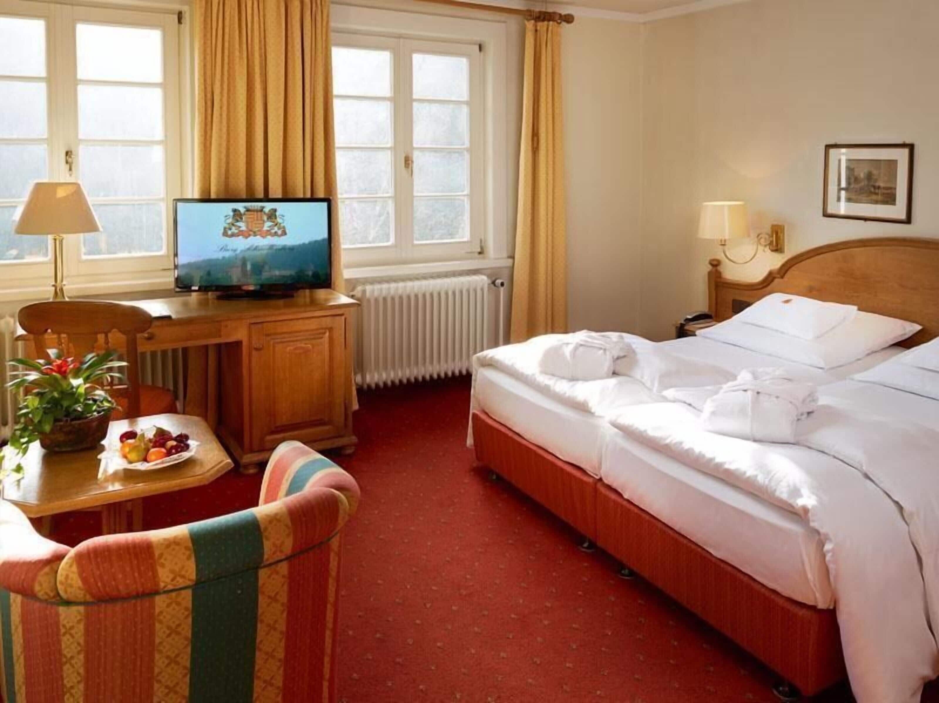 A hotel room with a bed, a desk with a television, a chair, a plant, and a window with curtains.