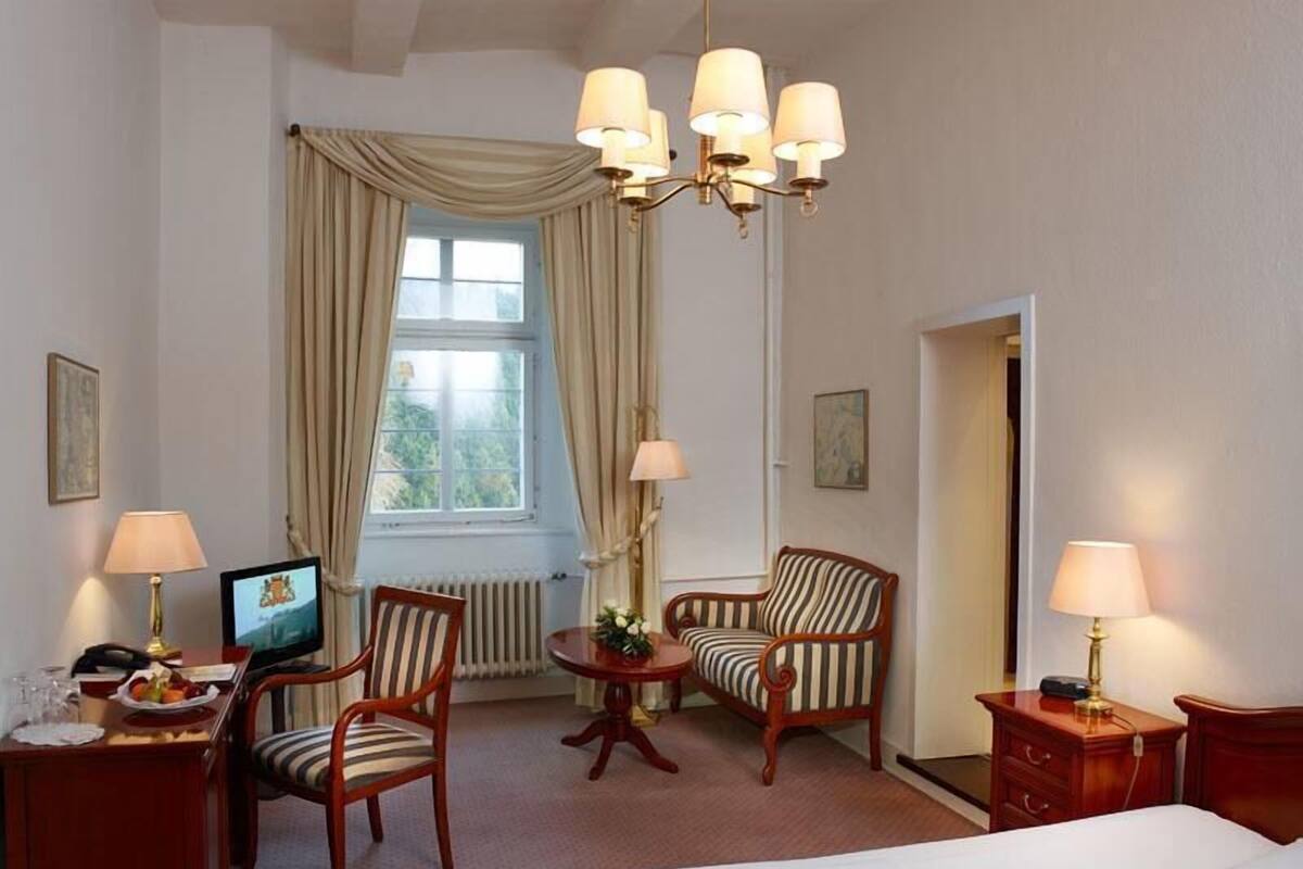 A hotel room with a bed, two chairs, a desk with a computer, a lamp, and a window with curtains.