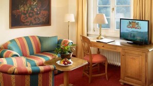 A cozy room with a striped sofa, a wooden desk with a computer, a chair, a small table with a plant and fruit, and a painting on the wall. - Burghotel Schnellenberg (Attendorn)