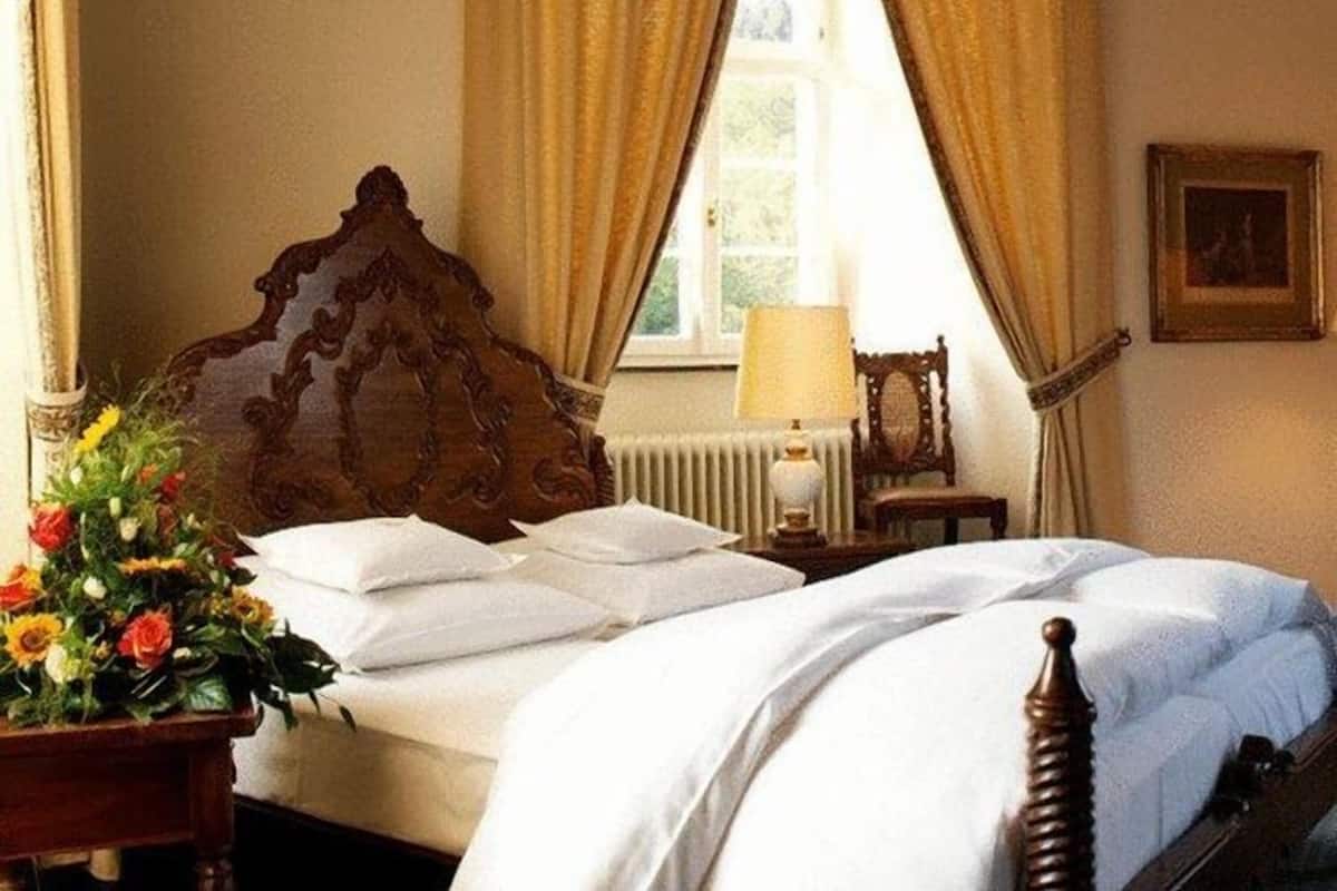 A bedroom with a wooden headboard, a bedside table with a lamp, a chair, and a window with curtains.