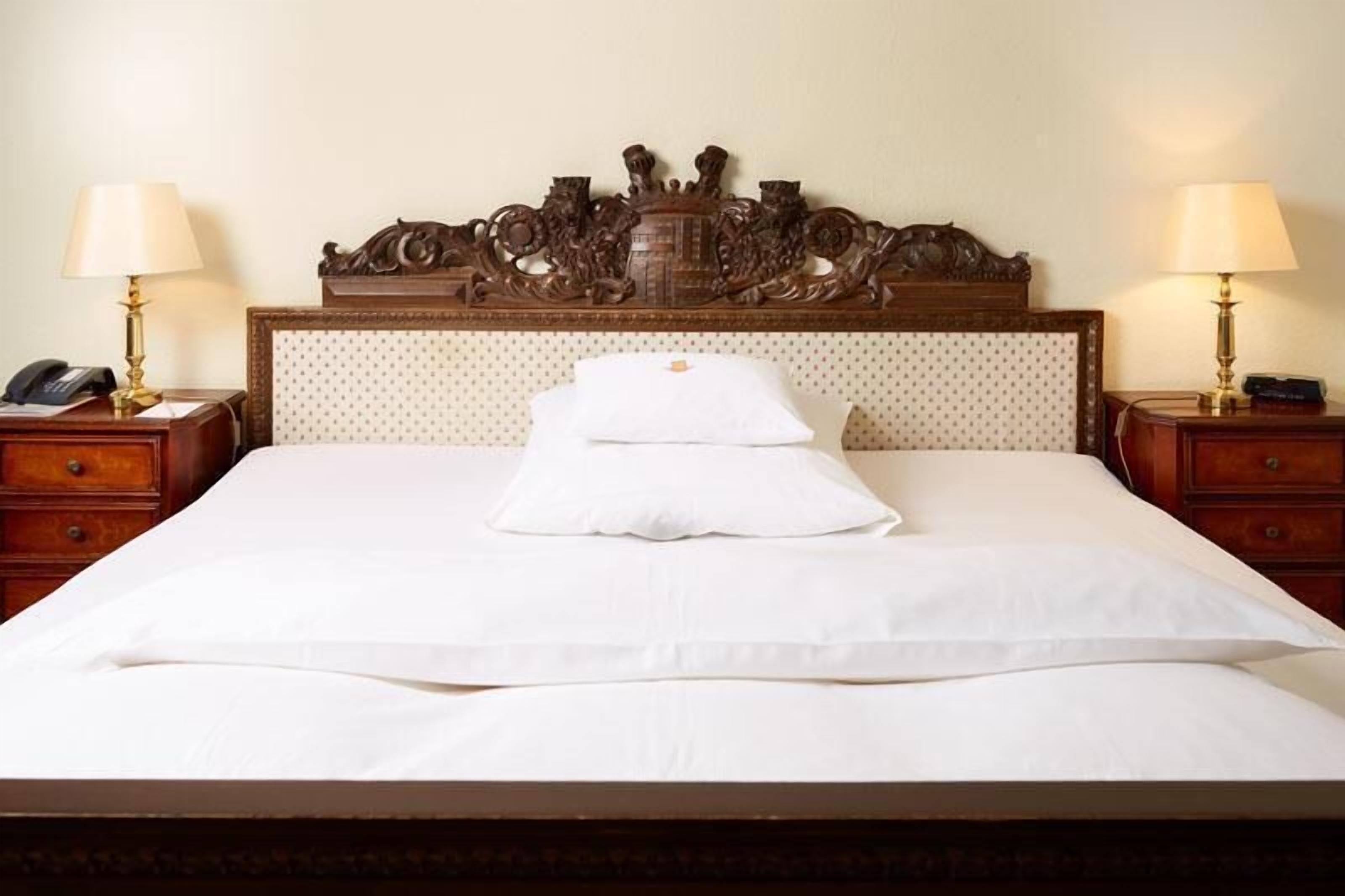 Ein Hotelzimmer mit einem großen Bett, zwei Nachttischen, einem verzierten Kopfteil und einem Telefon auf einem der Tische.