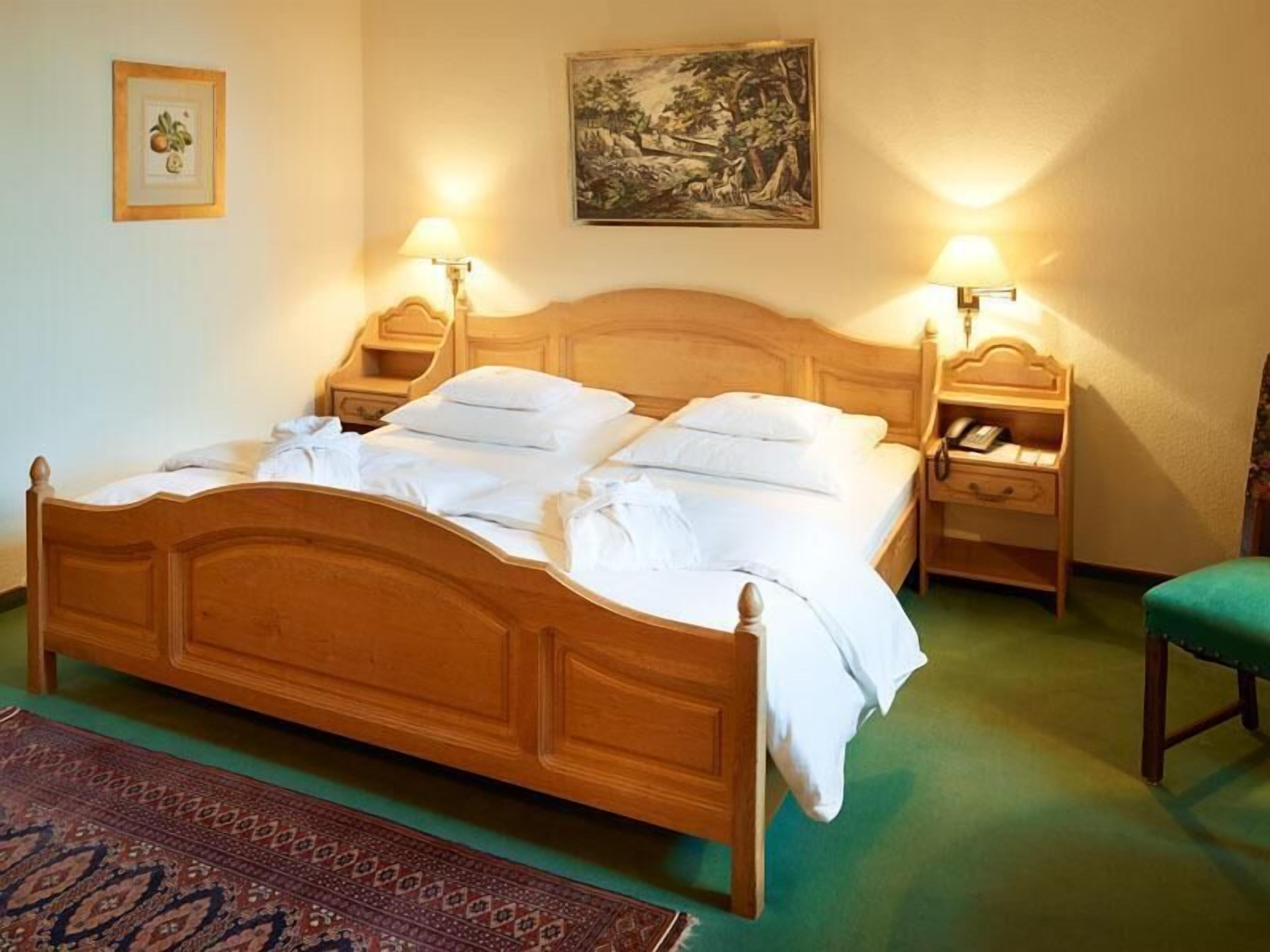 A wooden four-poster bed with white linens, two bedside tables with lamps, a framed botanical print, and a landscape painting on the wall.