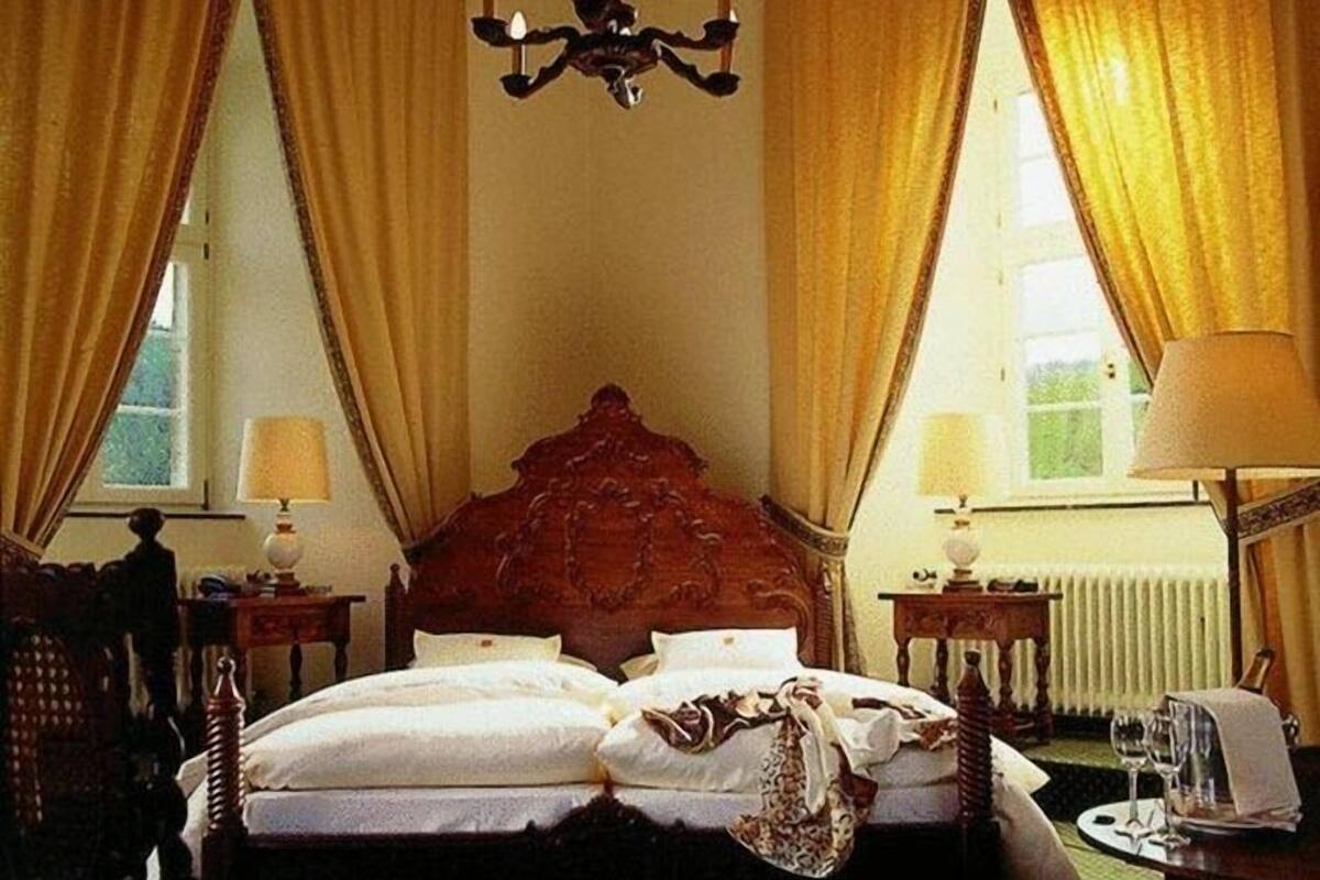 A hotel room with a large bed, two bedside tables with lamps, a chandelier, and curtains.