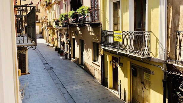 A narrow street lined with yellow buildings, each with balconies adorned with potted plants. - Il Terrazzino (Iglesias)