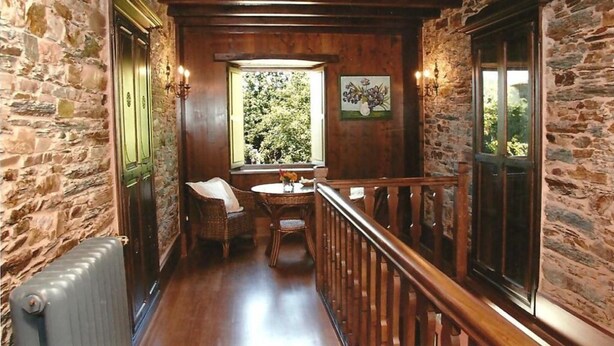 A narrow corridor with stone walls, wooden flooring, a wooden railing, a table with chairs, and a window with a view of greenery. - Casa Rural Vila Pomar (Ribadeo)