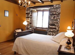 A bedroom with a bed, a nightstand, a lamp, a window with a view, and a stone wall. - Casa Rural Vila Pomar (Ribadeo)