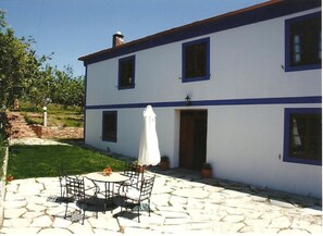 A two-story building with a white exterior and blue trim. There is a patio with stone flooring and a table with chairs. - Casa Rural Vila Pomar (Ribadeo)
