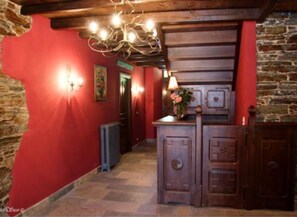 A narrow hallway with stone walls, a red accent wall, and a wooden ceiling. There is a chandelier, a door, and a small table with a vase of flowers. - Casa Rural Vila Pomar (Ribadeo)