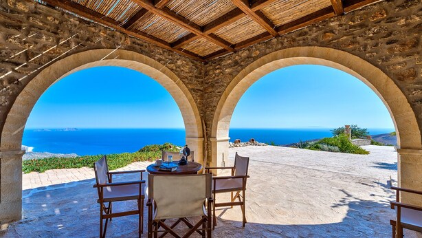 Eine Steinterrasse mit zwei Holzstühlen und einem Tisch, mit Blick auf das Meer.