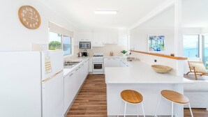 A modern kitchen with white cabinetry, a wooden floor, a large island with stools, a microwave, and a stove. - Ocean Dreaming - Brooms Head (Brooms Head)