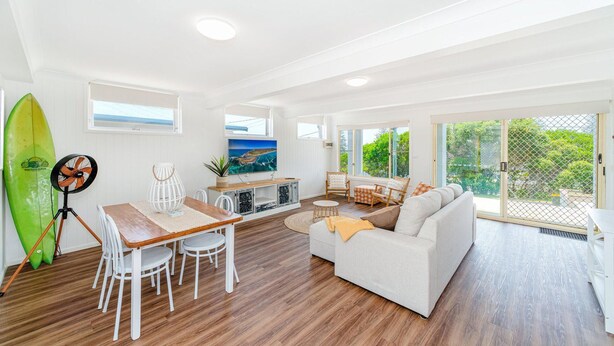 A spacious living area with a wooden floor, a white sofa, a dining table with chairs, and a surfboard against the wall. - Ocean Dreaming - Brooms Head (Brooms Head)