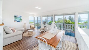 A spacious living room with a wooden dining table, white chairs, a sofa, and a balcony with a view of the ocean. - Ocean Dreaming - Brooms Head (Brooms Head)