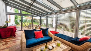 A covered patio with wicker furniture, a table with a bottle and candles, and a view of the garden.