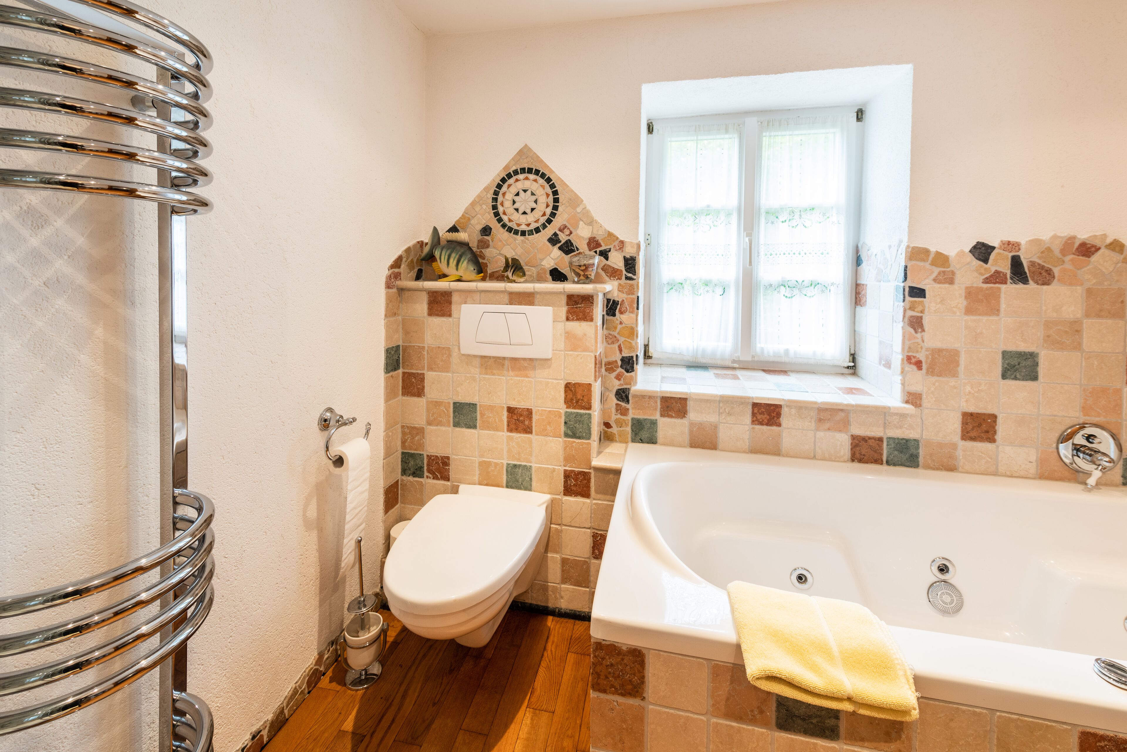 Une salle de bain avec un mur décoré d’une mosaïque, une baignoire, des toilettes, une fenêtre et un porte-serviettes.