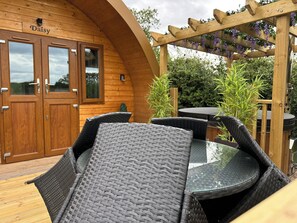 Una cabaña de madera con un letrero que dice "Daisy", con muebles de mimbre y una pérgola adornada con plantas colgantes.