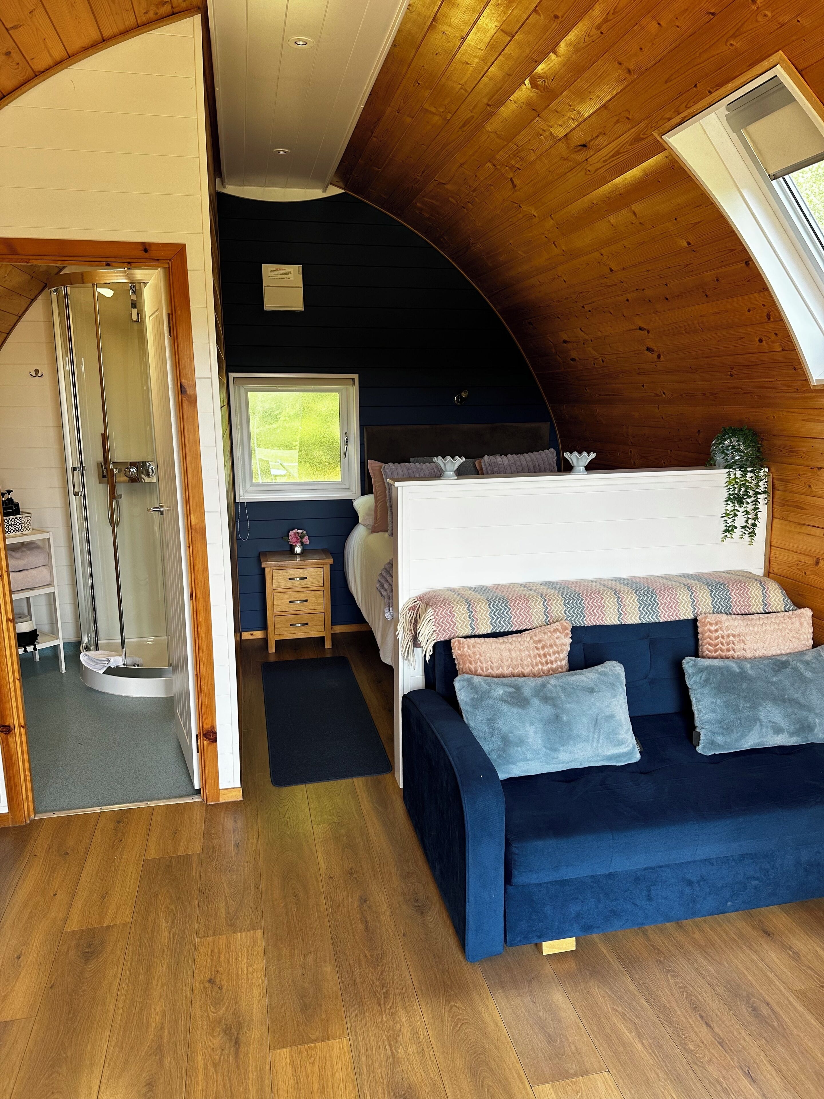 Un dormitorio con techo curvo, una cama con varias almohadas, una mesita de noche, una ventana y una zona de ducha.