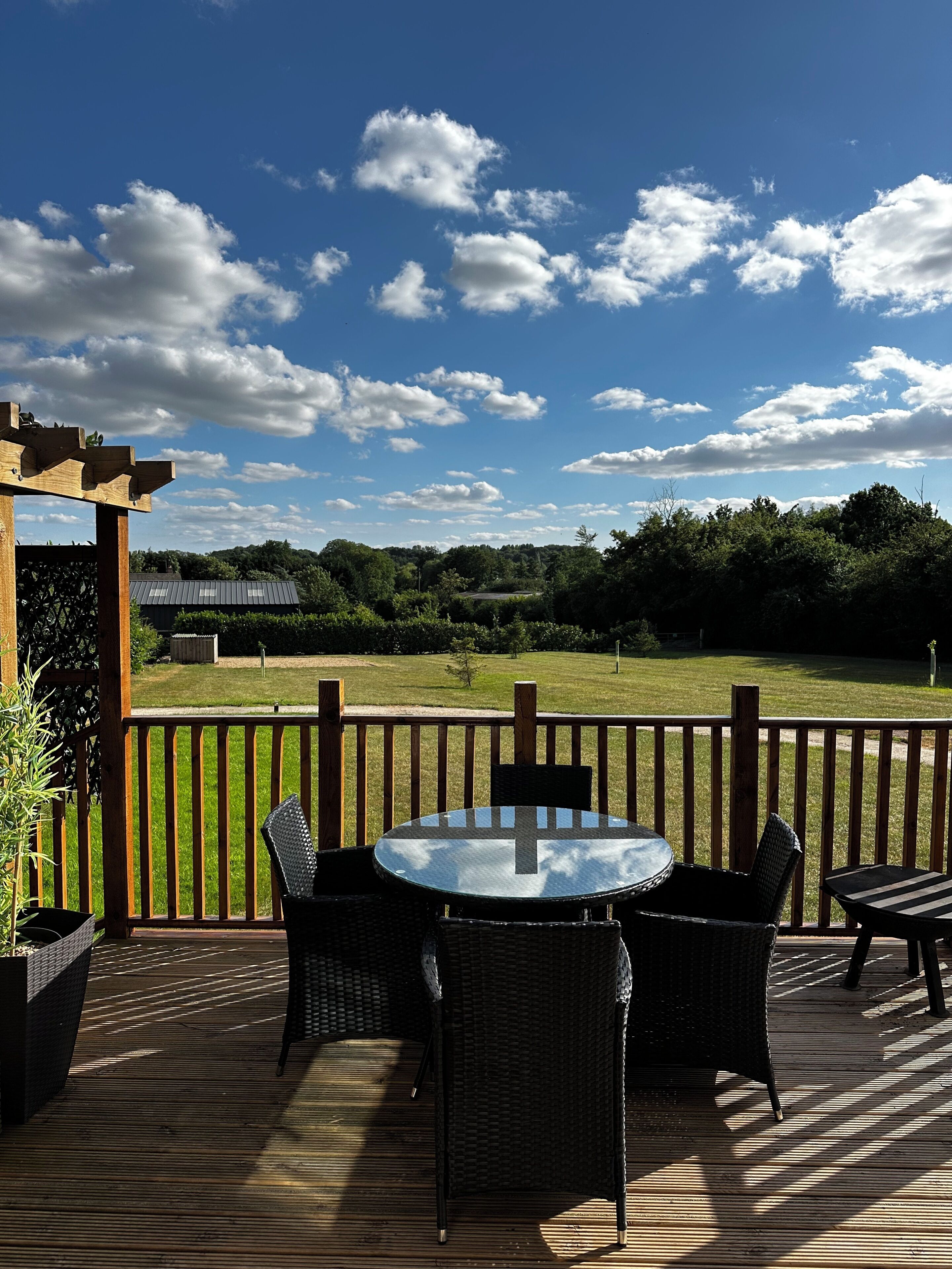 A wooden deck with a round table and chairs, a pergola, and a view of a grassy field under a partly cloudy sky.