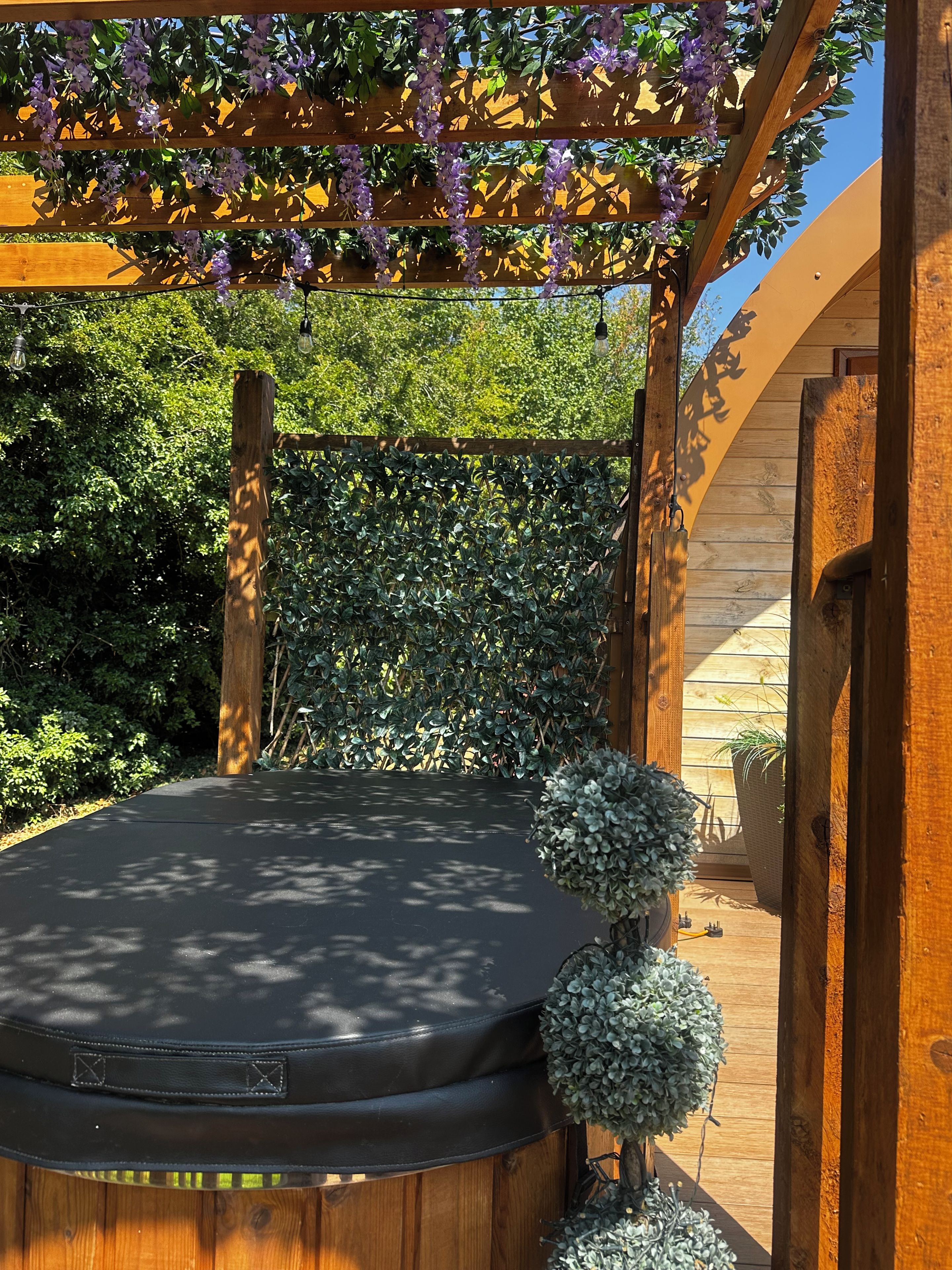 A wooden deck with a hot tub, surrounded by lush greenery and a pergola with purple flowers.