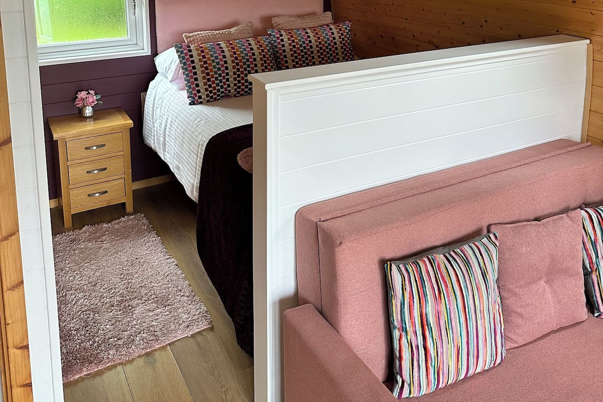 A cozy bedroom with a sloped ceiling, a bed with pink bedding, a bedside table with a lamp, and a window providing natural light.