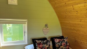 Un dormitorio con techo de madera curvada, una cama con almohadas florales, una mesita de noche con un florero y una ventana con persianas.