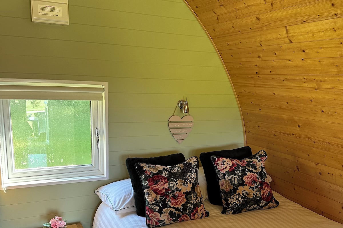 A bedroom with a curved wooden ceiling, a bed with floral pillows, a bedside table with a vase, and a window with blinds.