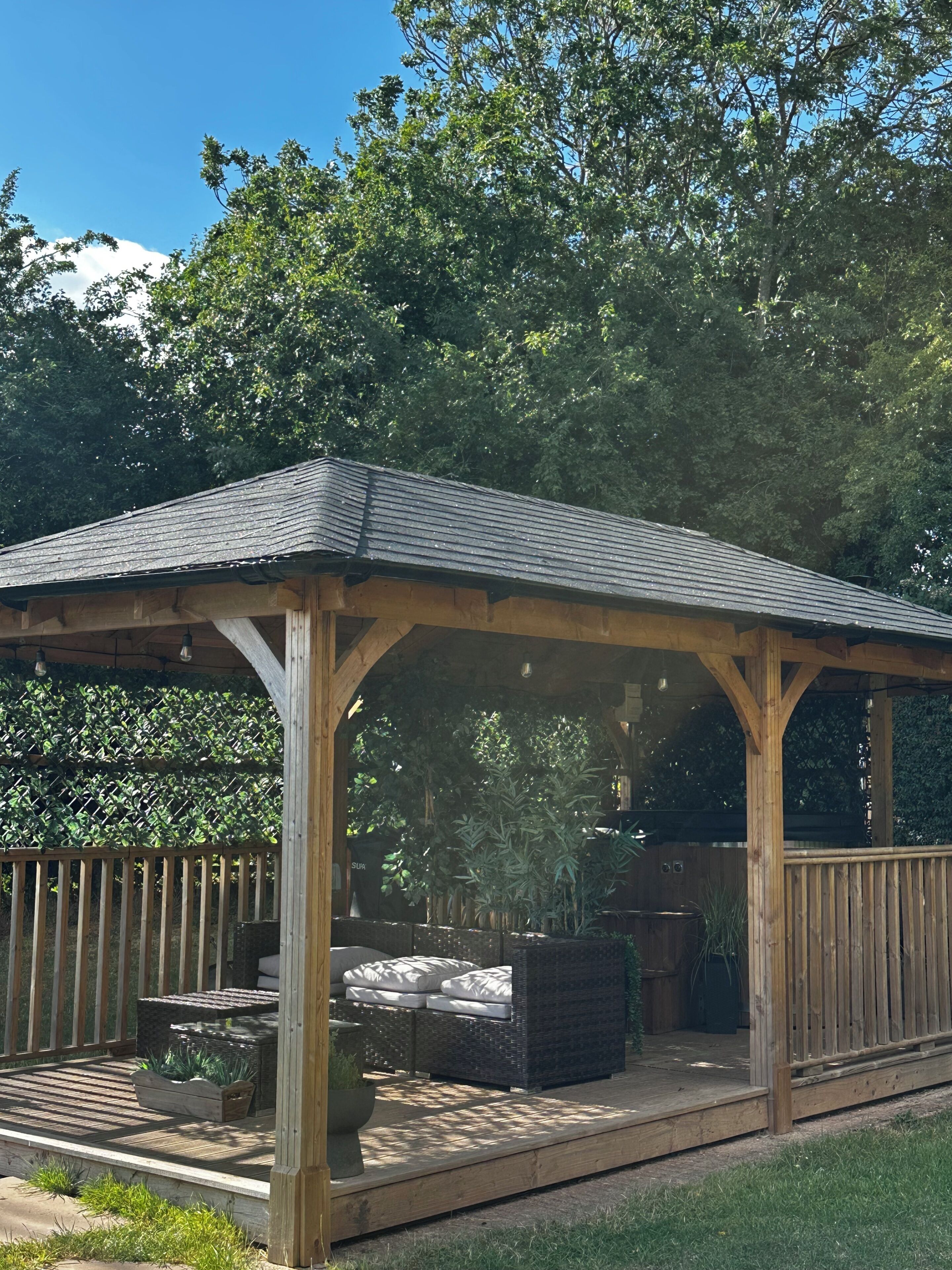 Ein hölzerner Gazebo mit geneigtem Dach, umgeben von einer Holzterrasse und einem Hecken.