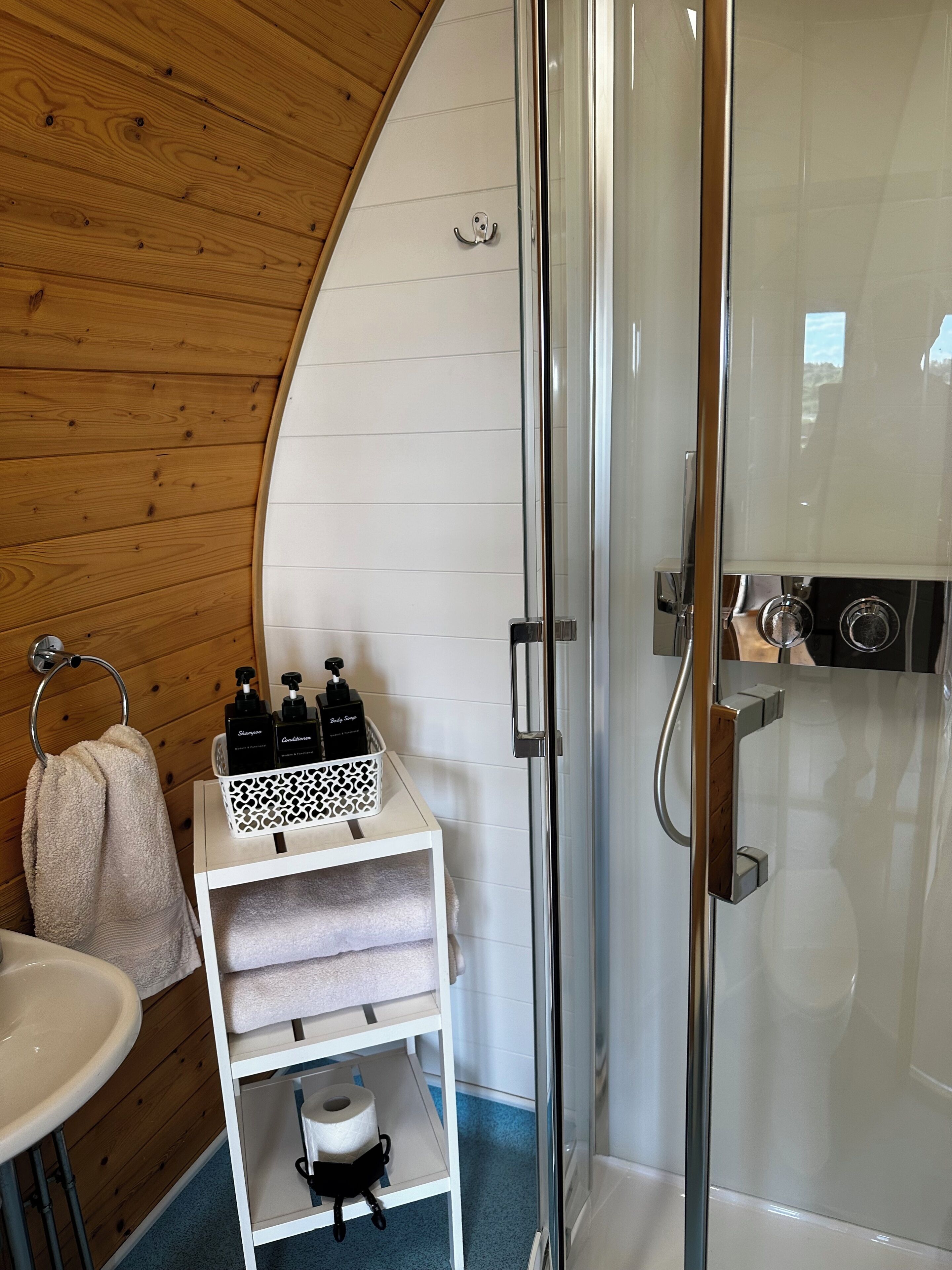 A bathroom with a glass shower enclosure, wooden wall paneling, a white sink, and a shelf with toiletries.
