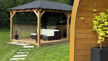 A wooden gazebo with a tiled roof, surrounded by a grassy lawn and a wooden fence.