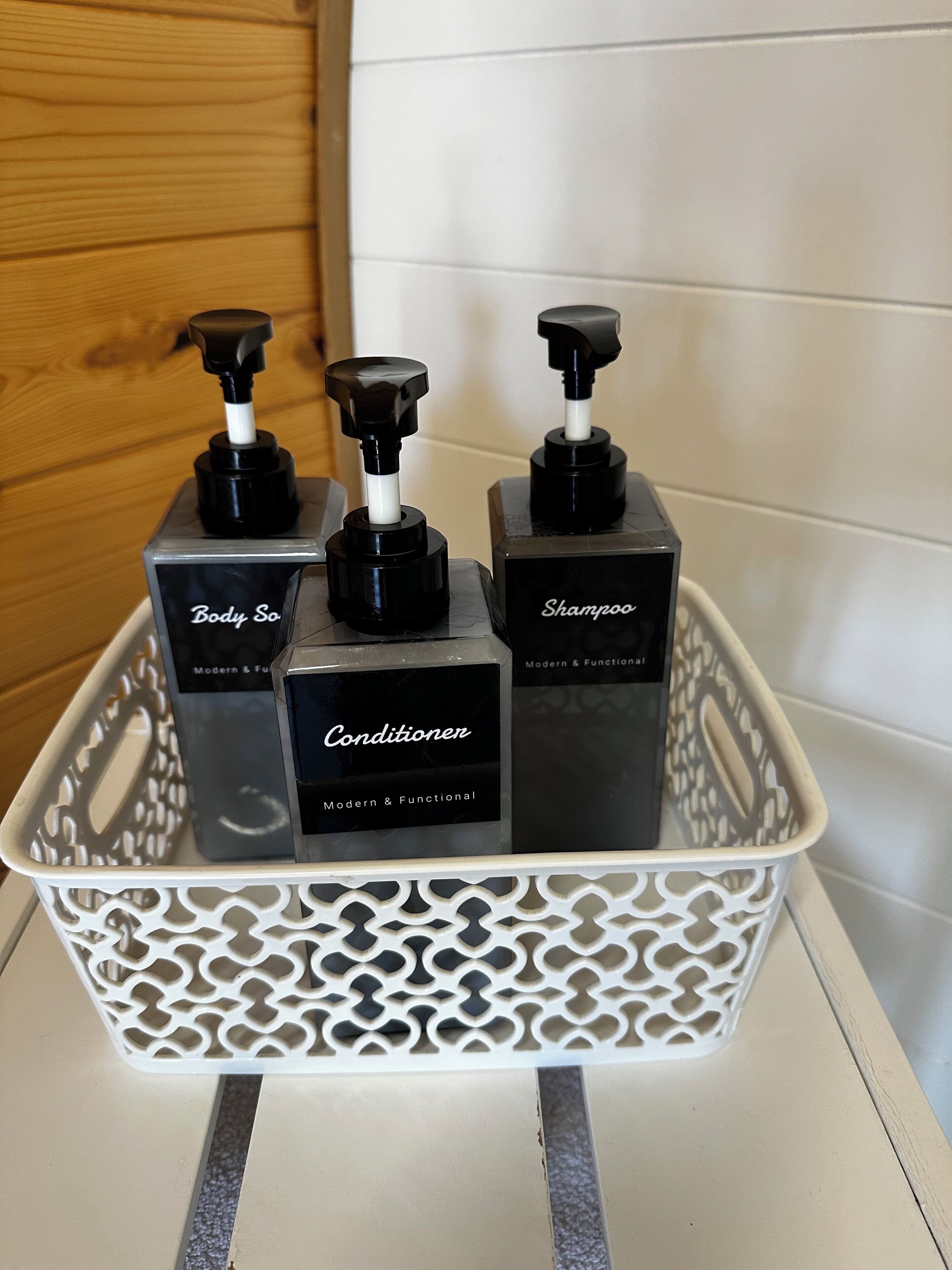 Ein Badezimmer mit weiß gefliesten Wänden und einer Holzverkleidung. Ein weißer Kunststoffkorb enthält vier schwarze Flaschen mit Duschgel, Conditioner und Shampoo.