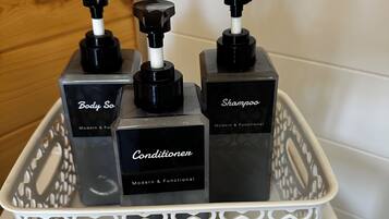 A bathroom with a white tiled wall and a wooden panel. There is a white plastic basket containing four black bottles of body soap, conditioner, and shampoo.