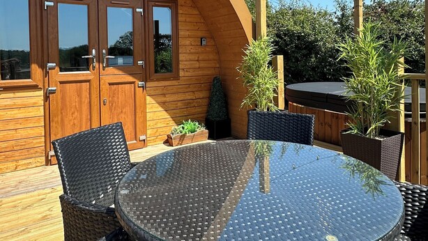 Una cabaña de madera con puerta de cristal que dice "Daisy", una mesa redonda de cristal, sillas de mimbre y plantas en macetas.