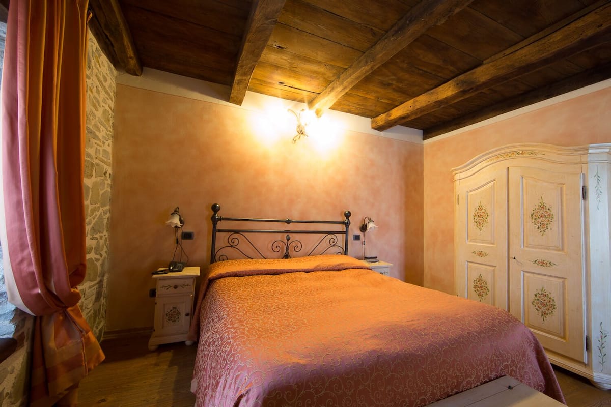 Ein Schlafzimmer mit Holzbalkendecke, einem Bett mit orangefarbener Bettdecke, einem Nachttisch mit Lampe, einem Schrank mit floralem Dekor und einer Steinwand.