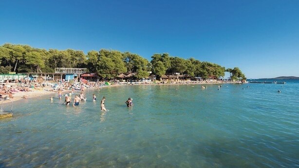 Una playa con agua cristalina, gente nadando y tomando el sol, y una hilera de árboles y edificios a lo largo de la orilla.