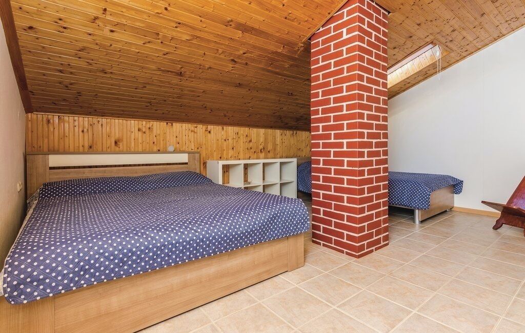A bedroom with a wooden ceiling, a brick chimney, a bed with a polka dot bedspread, and a small table with a chair.
