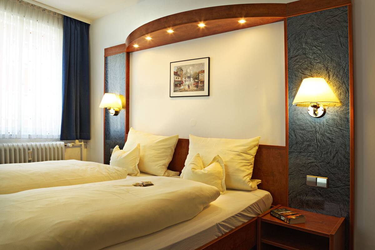 Een hotelkamer met twee bedden, een nachtkastje, een lamp, een lijst aan de muur en een raam met gordijnen.