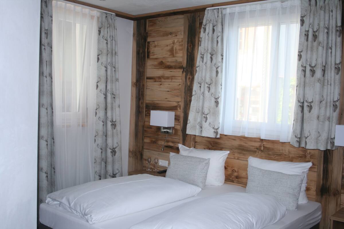 Een tweepersoonsbed met witte lakens, een houten hoofdbord en dunne gordijnen met hertenmotieven.