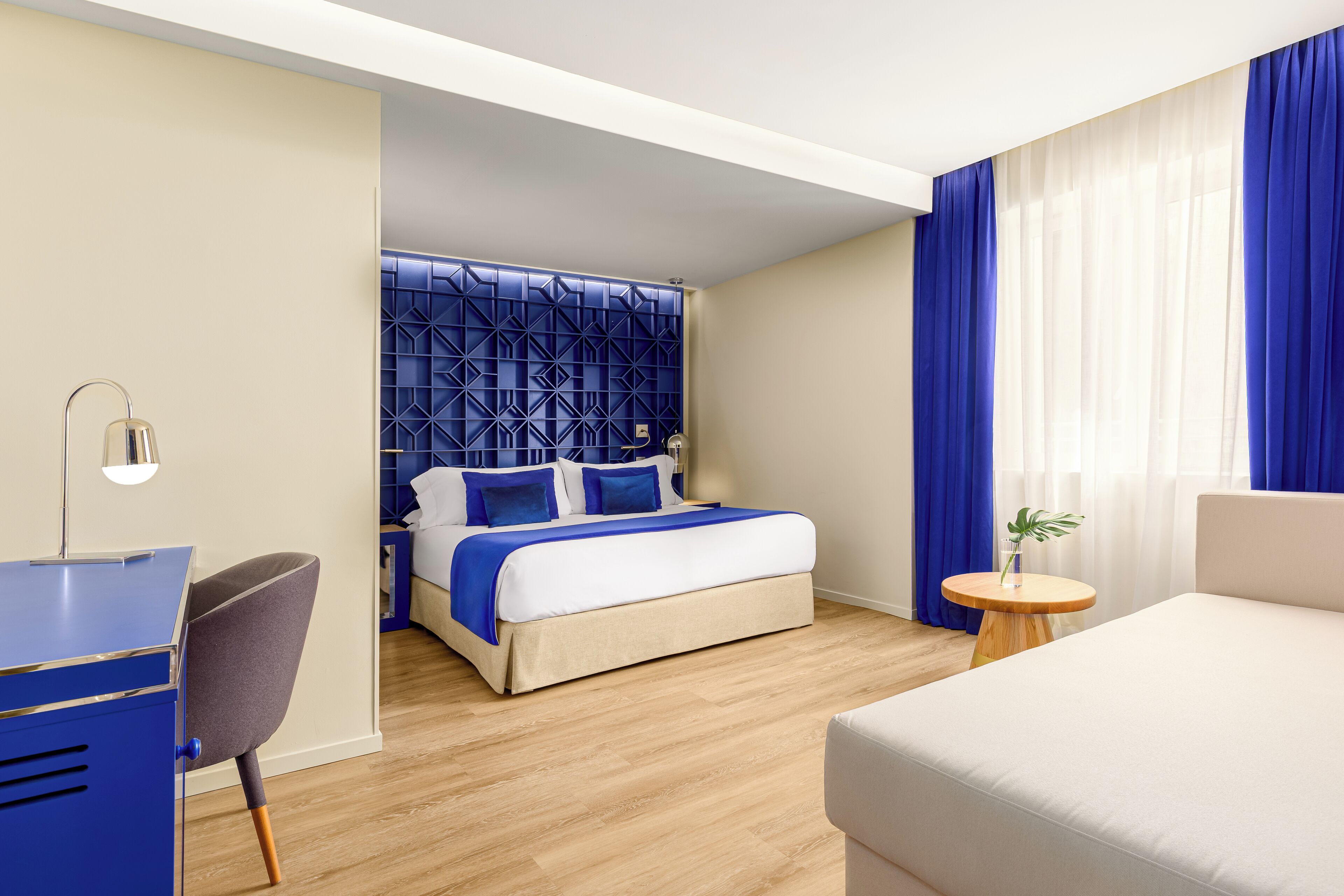 A modern hotel room with a bed, a desk, a chair, and a round wooden table.