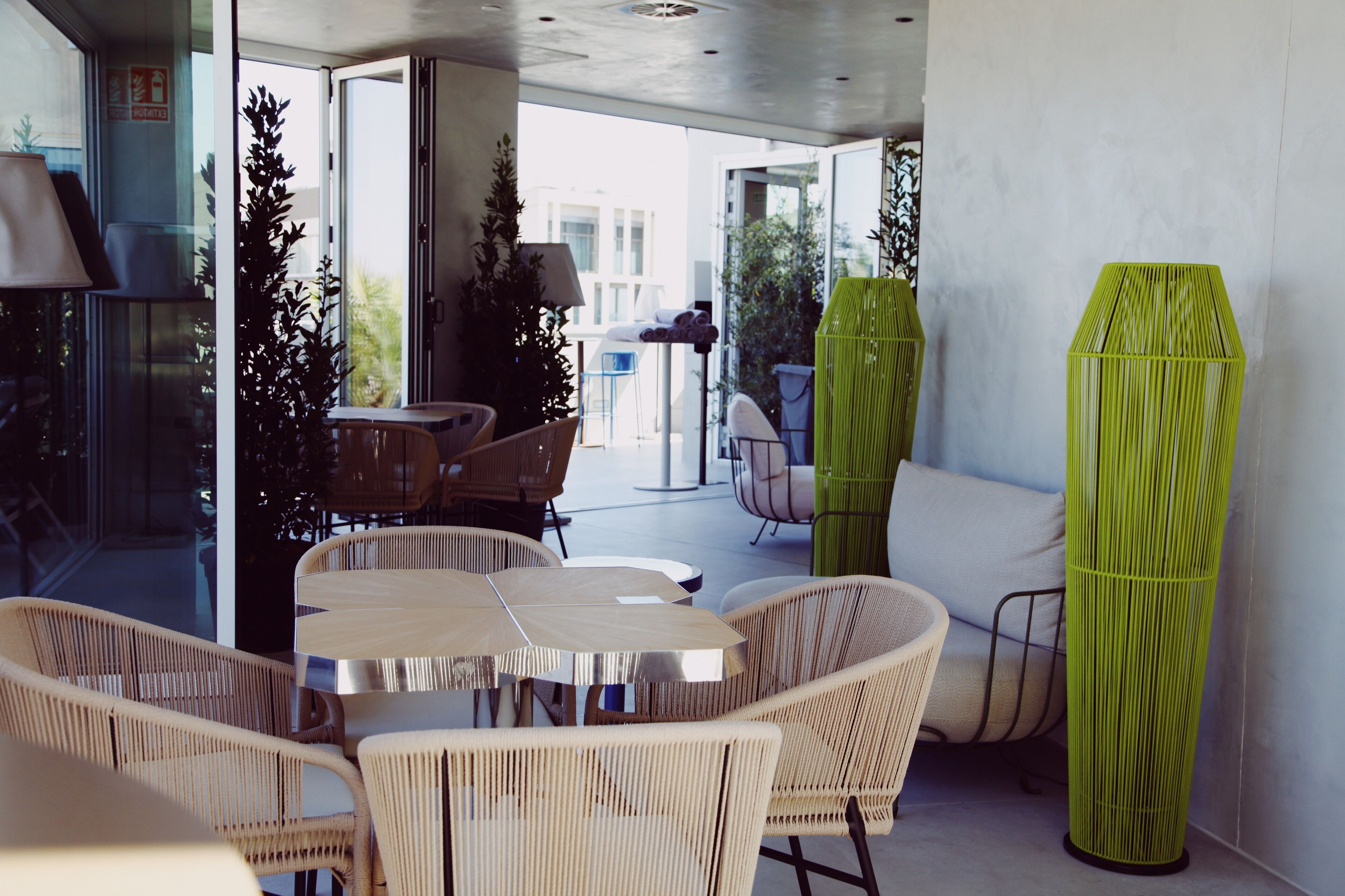 Un espacio exterior moderno con sillas de mimbre, una mesa redonda, puertas de cristal que dan a un balcón y una maceta cilíndrica verde.