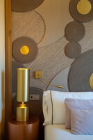 Una habitación moderna con pared texturizada, una lámpara dorada sobre una mesita de madera y una cama blanca con almohadas.