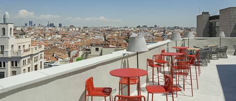 A rooftop terrace with red chairs and tables, offering a view of a cityscape with buildings and a clear sky.
