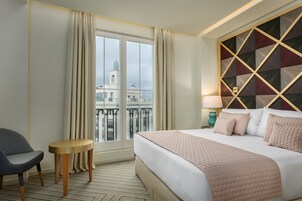 Camera d'albergo con un letto grande, una poltrona, un tavolino, vista su un edificio dalla finestra e un pannello decorativo al muro.
