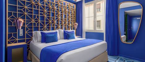 A modern hotel room with a geometric-patterned wall, a large bed with blue bedding, and a large mirror.