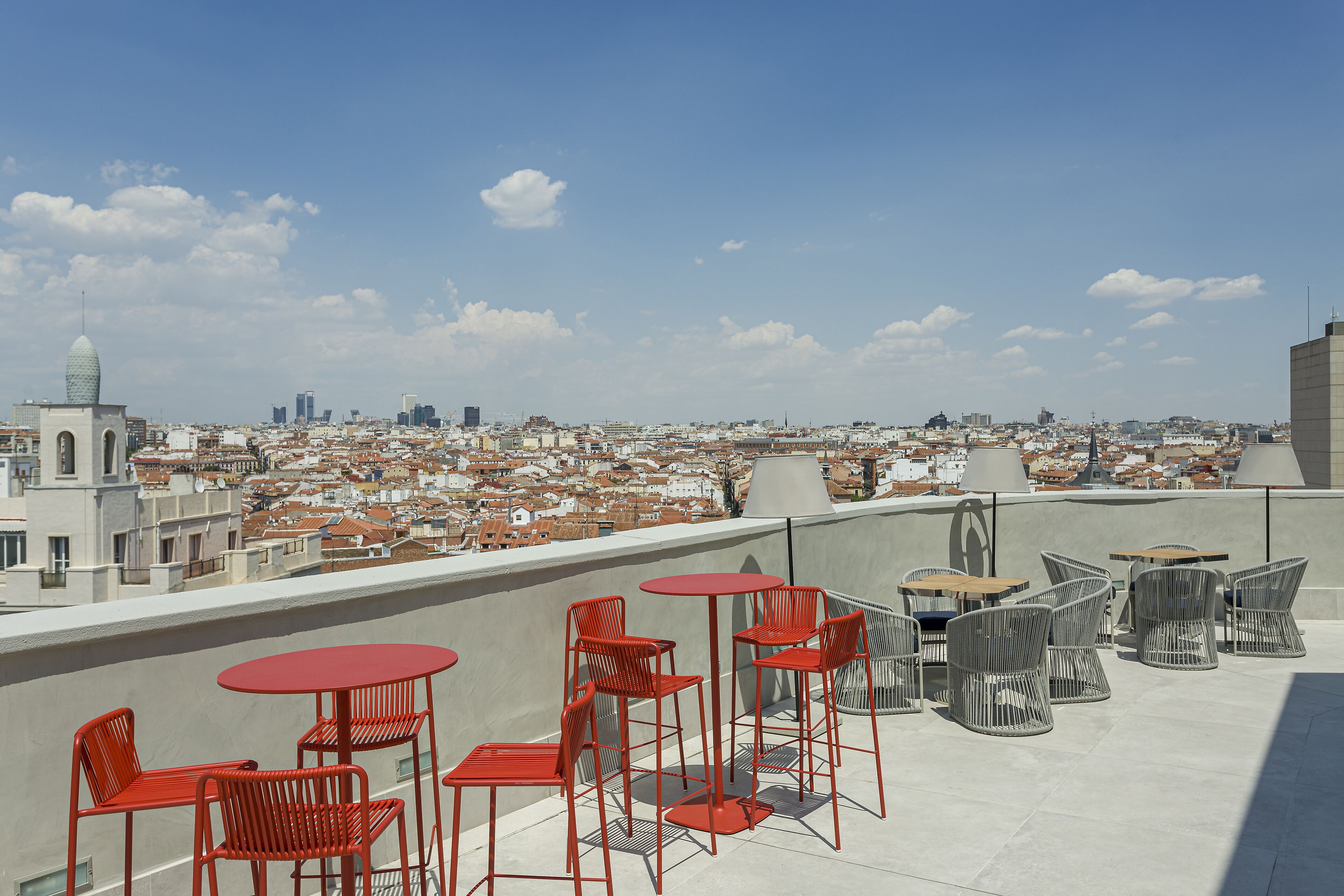 Une terrasse sur le toit, avec des chaises et des tables en métal rouge, offrant une vue panoramique sur un paysage urbain composé de différents bâtiments et d’un ciel dégagé.
