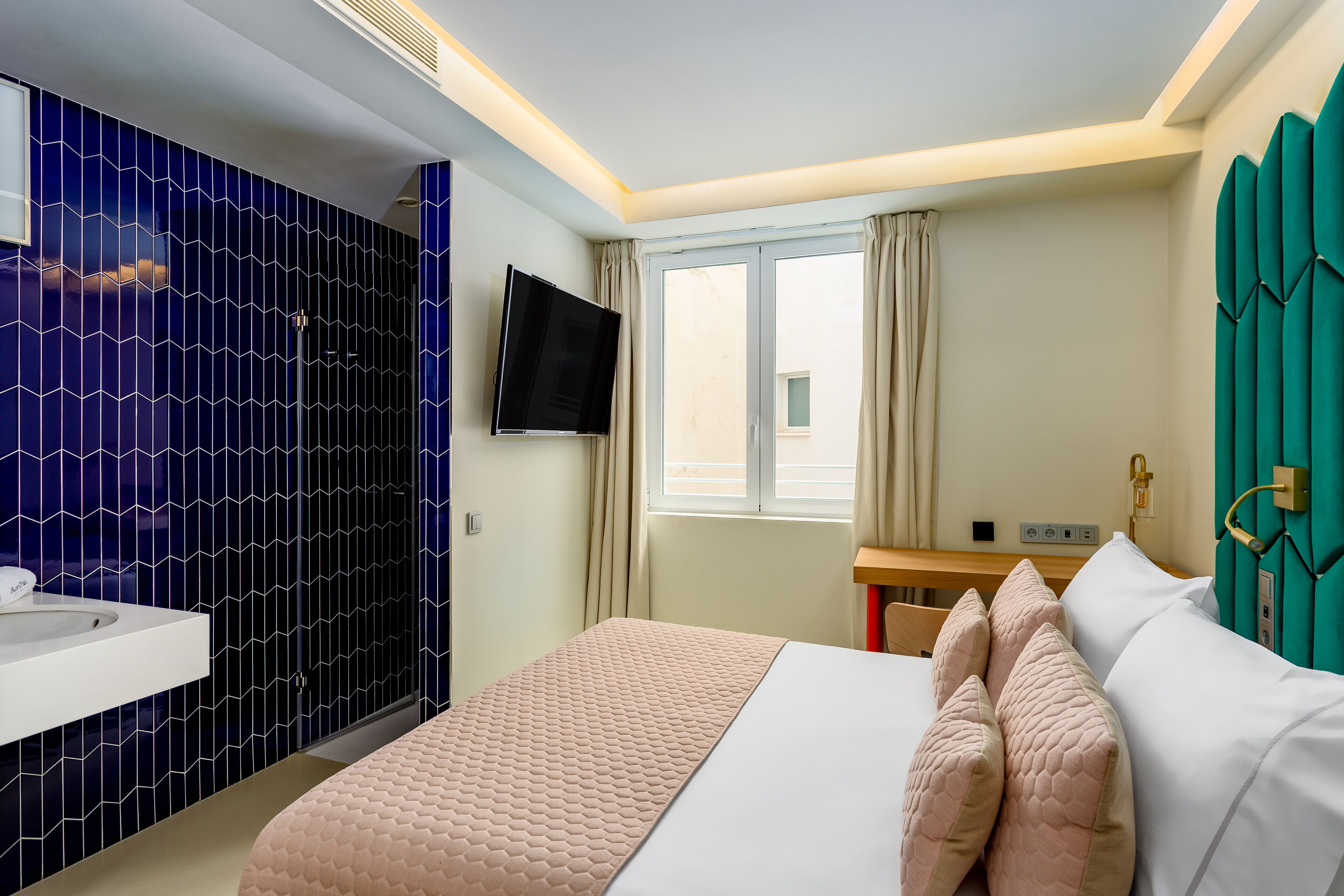 A hotel room with a bed, a TV, a window with curtains, a sink, and a teal-colored door.