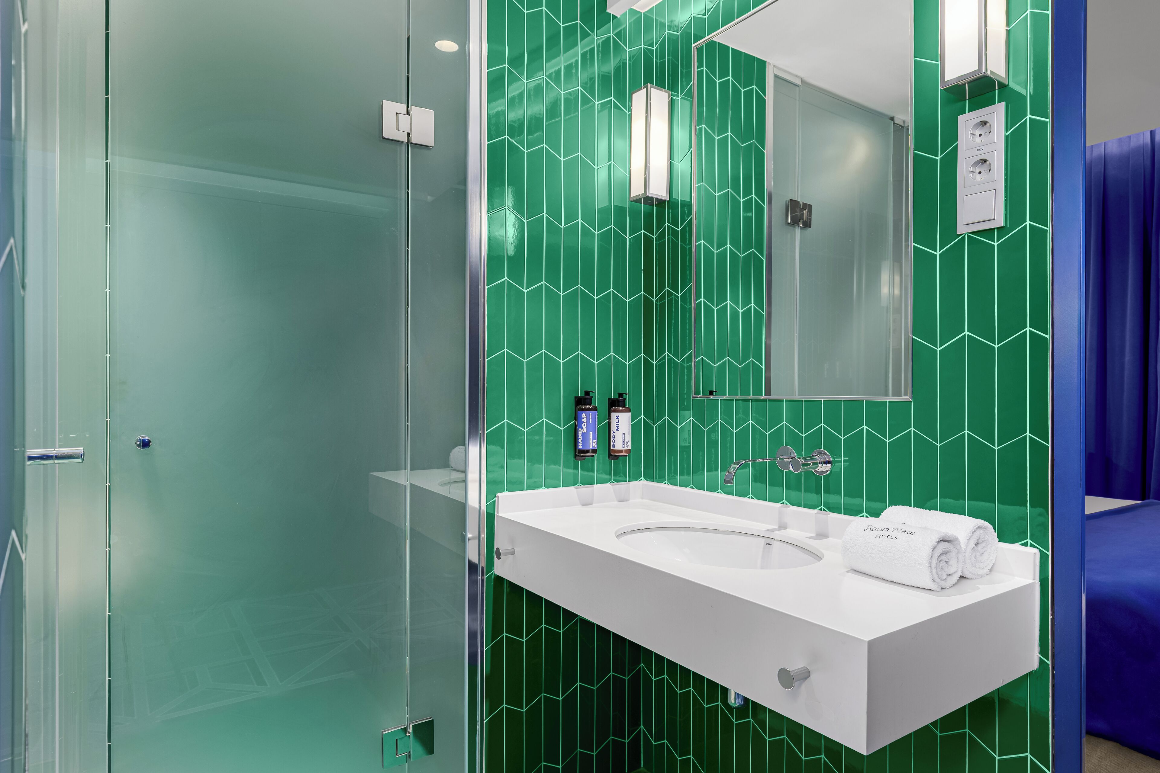 Ein modernes Badezimmer mit einer Duschkabine aus Glas, einem weißen Waschbecken mit Spiegel und grün gemusterten Fliesen.