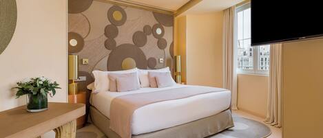 A modern hotel room with a large bed, a flat-screen TV, a decorative wall, and a view of buildings through the window.