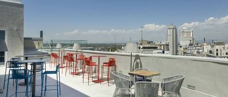 A rooftop terrace with outdoor seating, including tables and chairs, overlooking a cityscape.