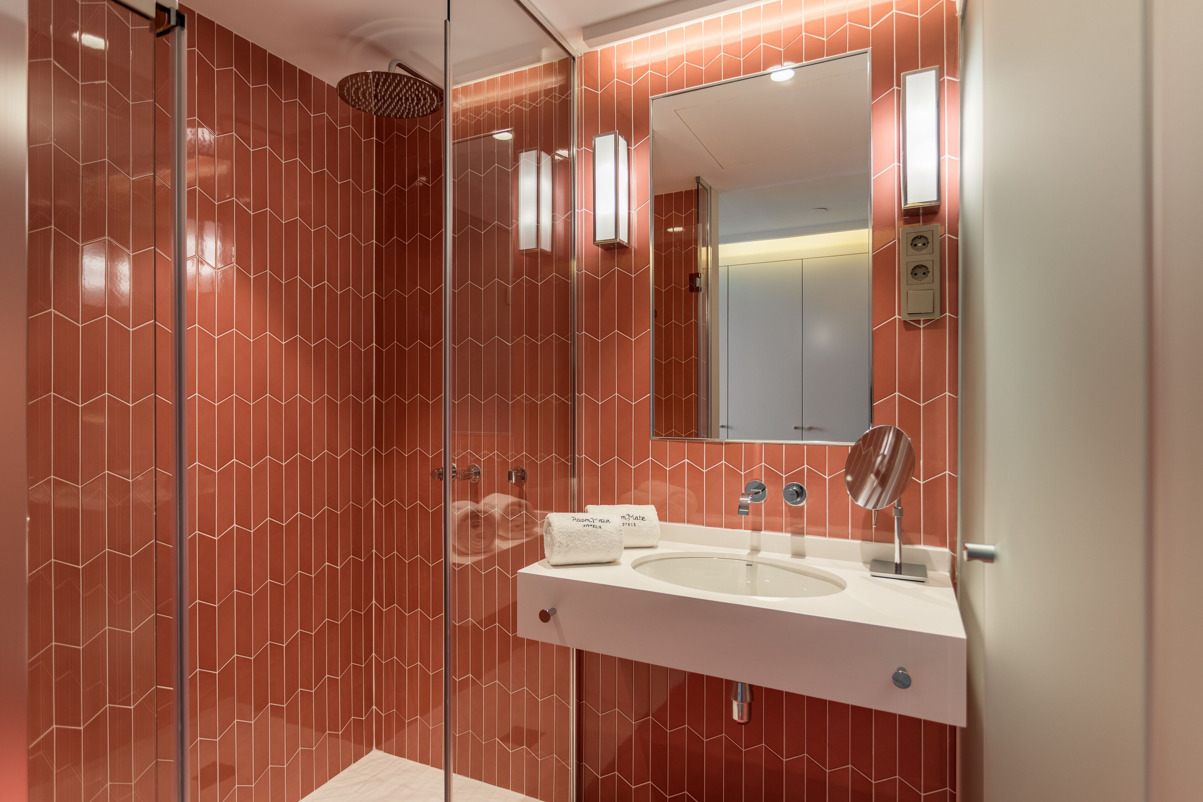 Une salle de bain moderne avec une douche à parois de verre, un lavabo blanc avec un miroir et un mur décoré d’un motif rouge.