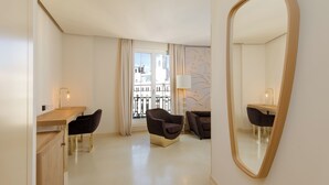 A hotel room with a wooden desk, two chairs, a large mirror, and a view of buildings through a window with curtains.