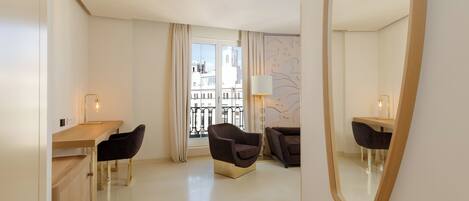 A hotel room with a wooden desk, two chairs, a large mirror, and a view of buildings through a window with curtains.